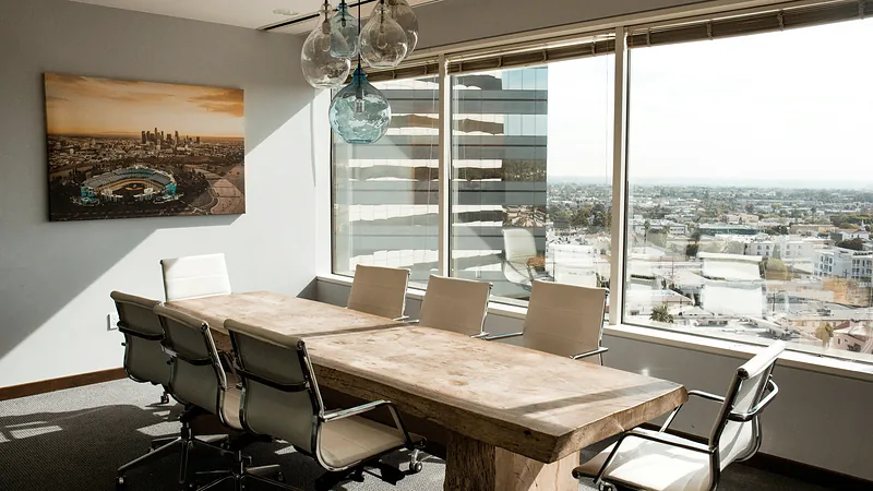 An empty wooden meeting table with chairs beside sunlit windows.