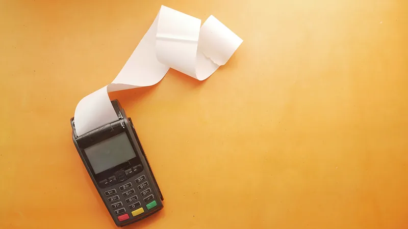 A payment terminal with a curling blank receipt on a warm yellow surface.