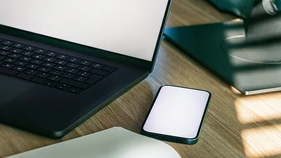 A laptop and phone with blank screens on a sunlit wooden desk.